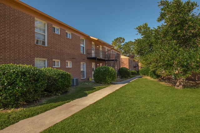 Exterior view of brick apartment buildings with manicured bushes and a concrete walkway.