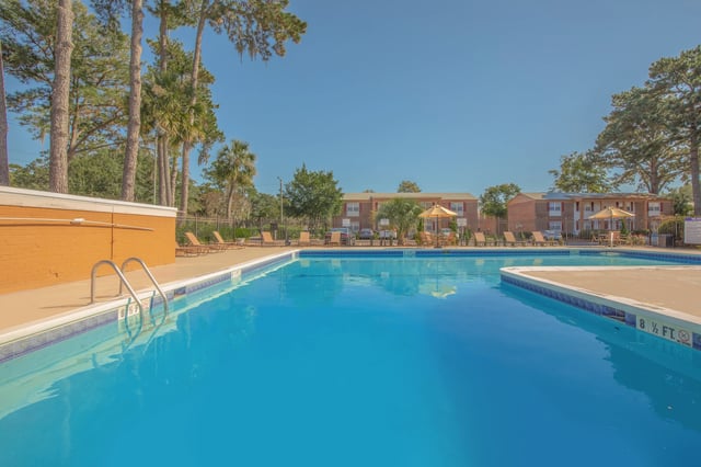 Outdoor swimming pool with lounge chairs and umbrellas, surrounded by trees and apartment buildings.