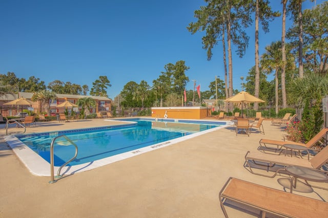 Outdoor swimming pool with lounge chairs and umbrellas, surrounded by trees and apartment buildings.