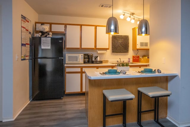 Kitchen area with black refrigerator, microwave, coffee maker, white cabinets, and a breakfast bar with two stools.