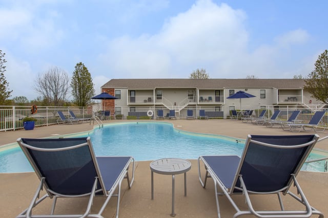 Outdoor swimming pool with lounge chairs and tables, with apartment buildings in the background.