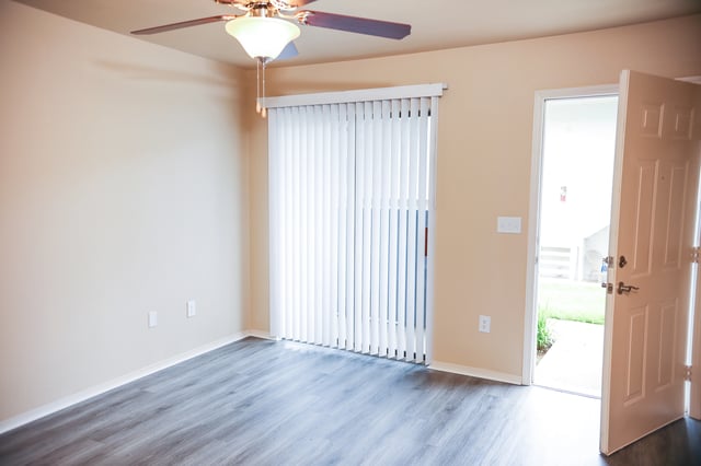 Interior view of an empty apartment room with beige walls, wood-look flooring, a ceiling fan, and an open front door.