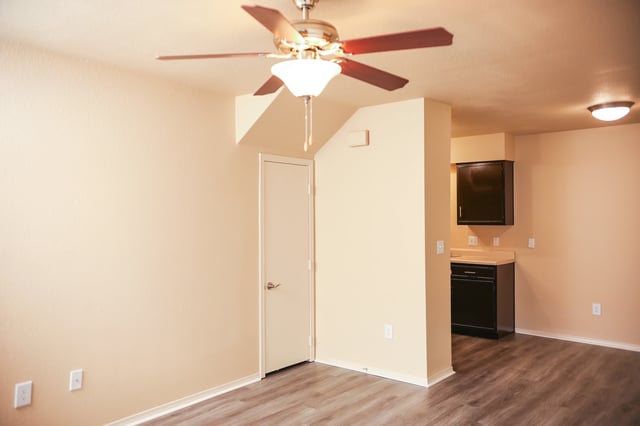 Interior of a living room with a ceiling fan and a view into a kitchen area.