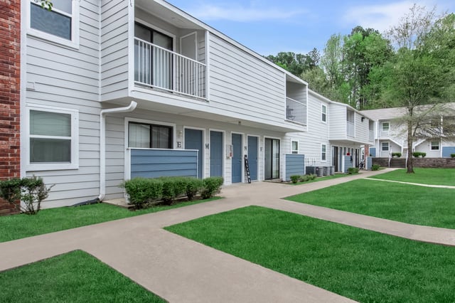 Exterior view of apartment buildings with blue doors and balconies, surrounded by green grass and trees.
