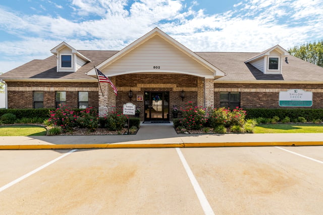 Shiloh Creek apartment complex leasing office with brick exterior, manicured bushes, and blooming pink flowers.