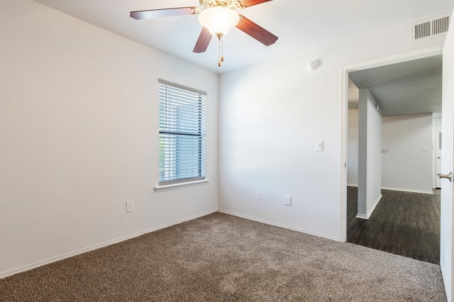 Interior view of a vacant apartment bedroom with plush carpeting, a ceiling fan, and a window with blinds.
