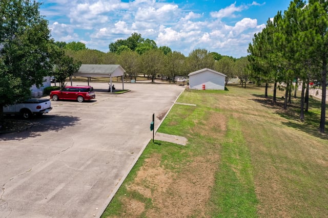Exterior view of parking lot with covered spaces, a white storage building, and lush green trees under a partly cloudy sky.