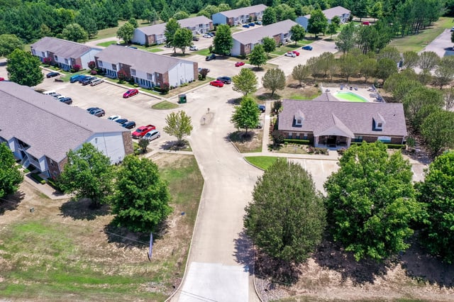 Aerial view of an apartment complex with multiple buildings, a swimming pool, and surrounding trees.