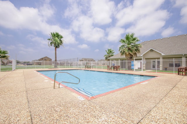 Outdoor swimming pool with lounge chairs and tables, surrounded by a white fence and palm trees.