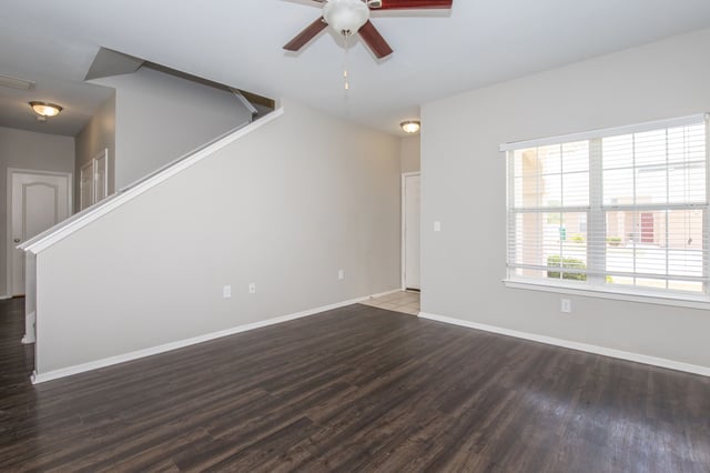 Interior view of a vacant apartment living room with dark wood laminate flooring, a staircase, and a large window.