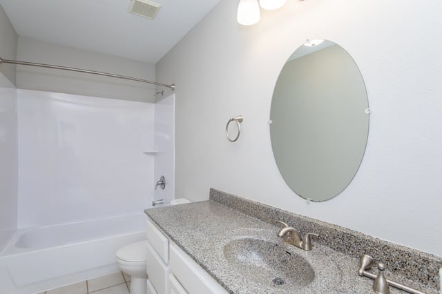 Bathroom with a granite countertop vanity, oval mirror, and white bathtub and toilet.