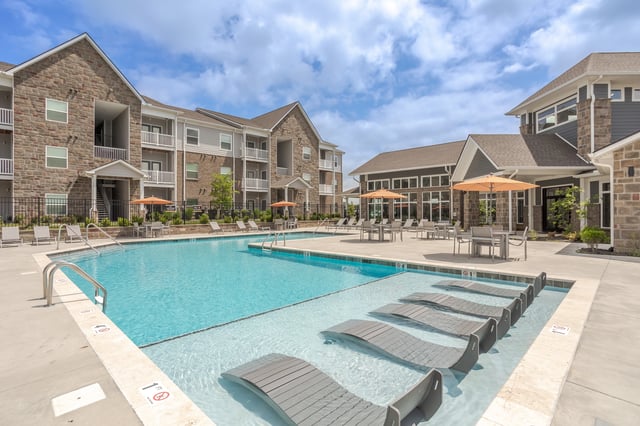 Outdoor swimming pool with lounge chairs and tables, surrounded by apartment buildings.