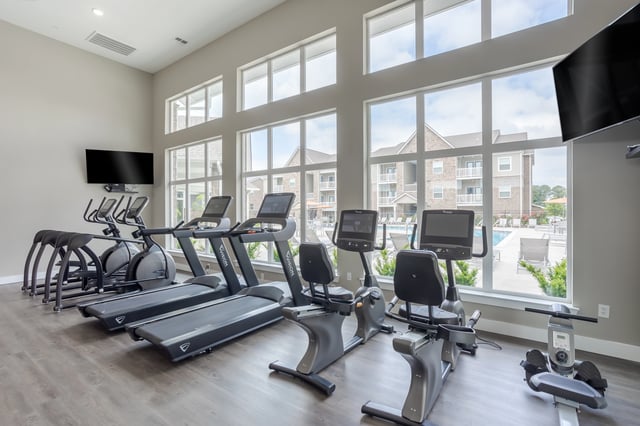 Treadmills and exercise bikes in a modern gym with large windows overlooking a pool and apartment buildings.