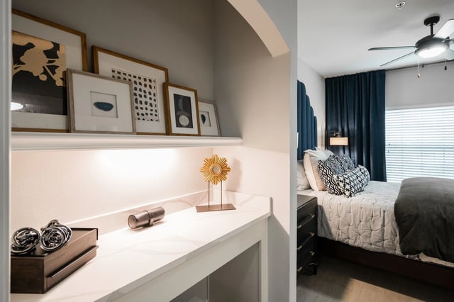 A chic built-in vanity area with framed art above, next to a cozy bedroom with a queen bed and navy blue curtains.