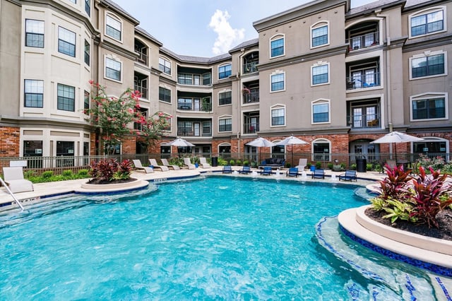 Resort-style swimming pool with lounge chairs and umbrellas, surrounded by apartment buildings.