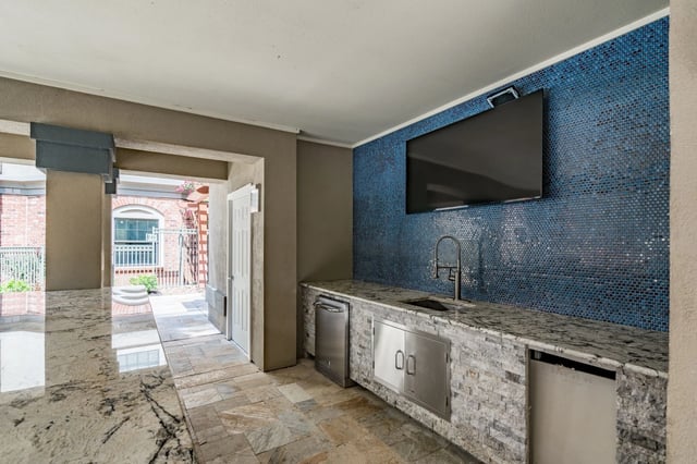 Outdoor kitchen with granite countertops, stainless steel appliances, and a blue mosaic tile backsplash with a TV mounted above.