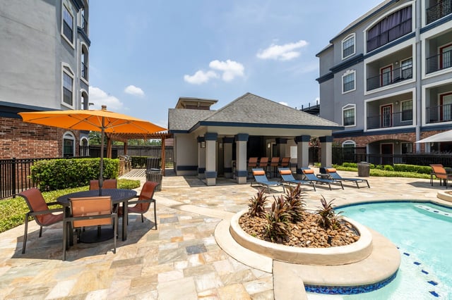 Outdoor swimming pool area with lounge chairs, tables, umbrellas, and a cabana.