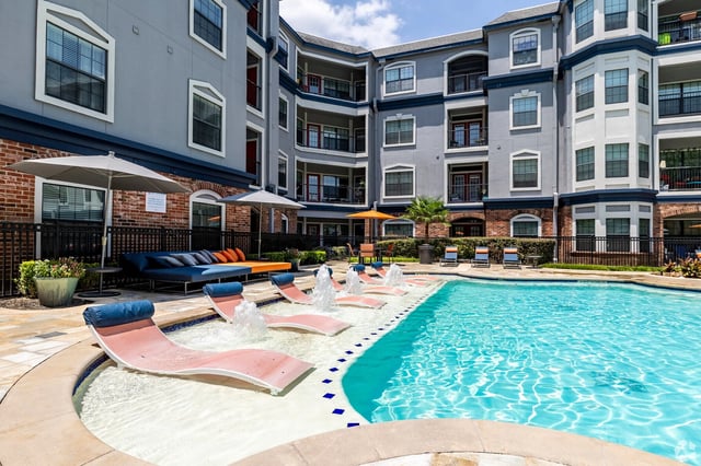 Outdoor swimming pool with tanning ledge and fountains, adjacent to apartment building.