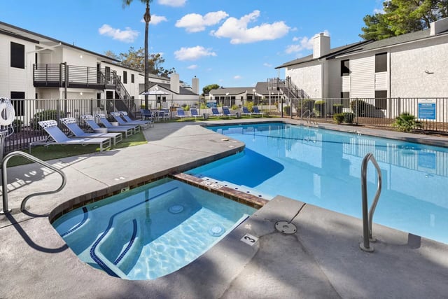 Swimming pool and jacuzzi area with lounge chairs and apartment buildings in the background.