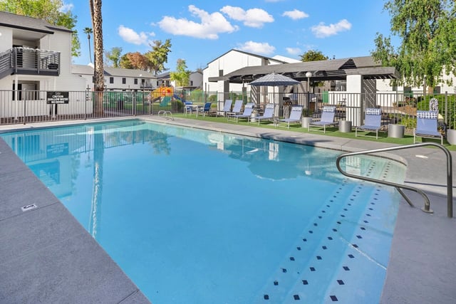 Bright blue swimming pool with lounge chairs and umbrellas surrounding it. Buildings and a playground are visible in the background.
