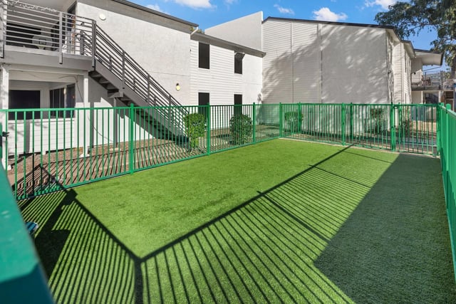Fenced-in artificial turf dog park with outdoor stairs and building exterior.