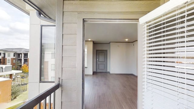 View from balcony into a spacious living room with a gray door and blinds on the window.