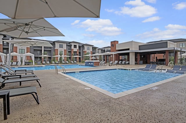 View of a resort-style swimming pool with lounge chairs and cabanas, adjacent to apartment buildings.