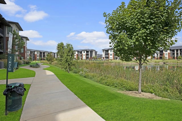 A paved walking path curves through a grassy area with a pond and apartment buildings in the background.