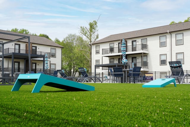 Outdoor courtyard with lounge chairs, tables, umbrellas, and cornhole boards in front of apartment buildings.