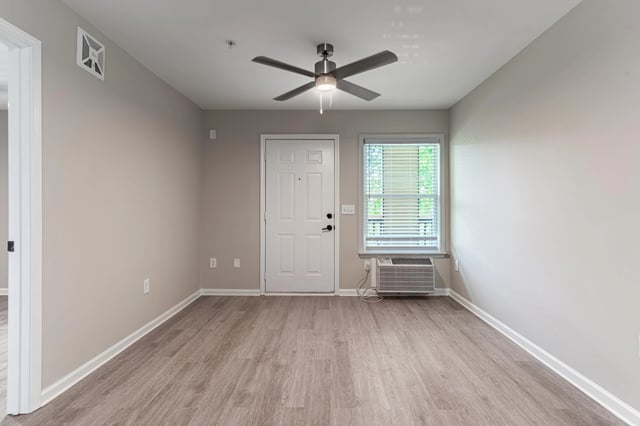 Empty living area with a ceiling fan, front door, and window with blinds.