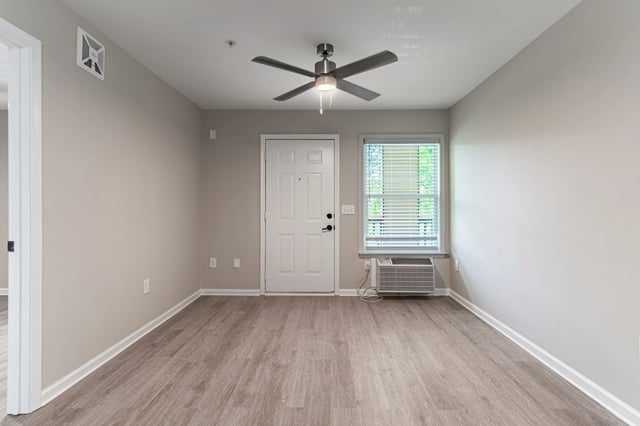 Empty living room with wood-look flooring, a ceiling fan, a white door, and a window with blinds.
