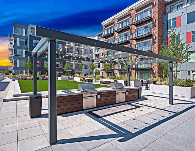 Outdoor grilling area with two built-in grills and string lights under a pergola, overlooking a courtyard with green space and apartment buildings.