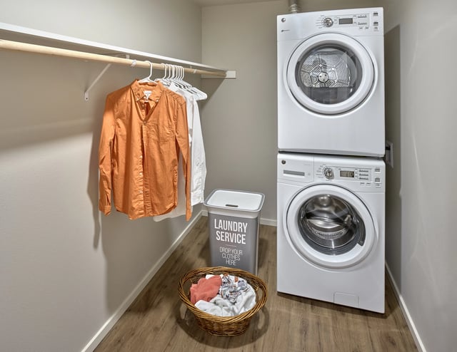Stacked white washer and dryer with a laundry hamper and clothes on hangers in a closet.
