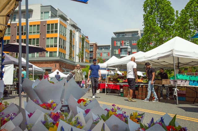 People browsing stalls at an outdoor farmer's market with modern apartment buildings in the background.