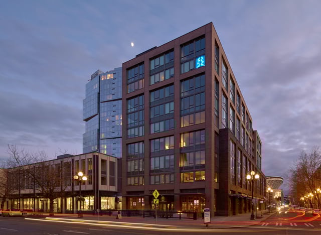 Exterior view of a modern apartment building at dusk with a neon blue logo on the side and traffic streaks on the street.