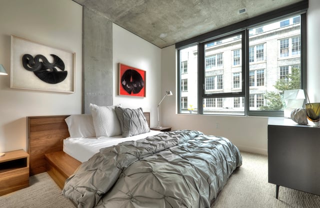 Modern bedroom with a king-size bed, concrete ceiling, and large windows overlooking a city street.