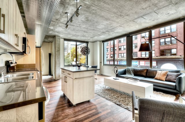 Modern living room and kitchen area with large windows, industrial ceiling, and sleek furnishings.