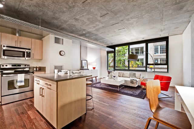 Modern studio apartment with open-concept kitchen and living area, featuring concrete ceilings, hardwood floors, and large windows.