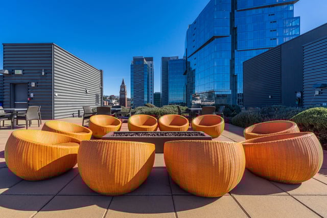 Rooftop lounge area with orange wicker chairs and a fire pit, overlooking city skyscrapers.
