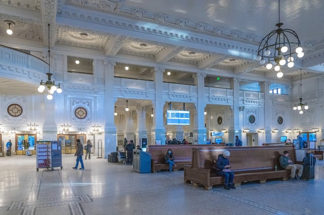 Interior of a grand train station waiting area with ornate architecture, seating, and departure boards.