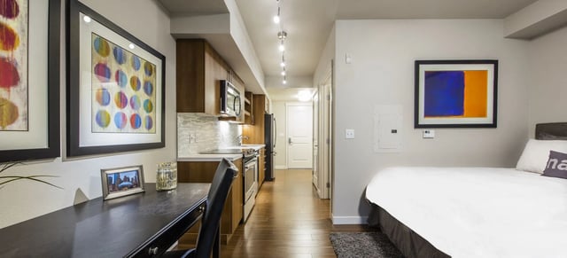 A modern studio apartment featuring a bed, desk, kitchen, and hallway.