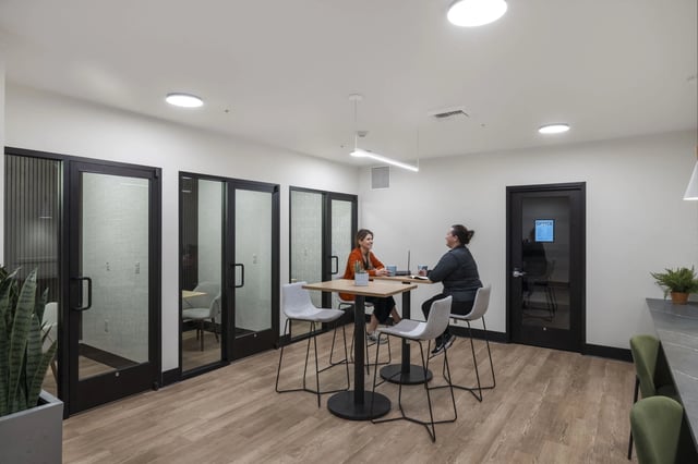 Two women collaborate at a high-top table in a shared office space with glass-walled meeting rooms.