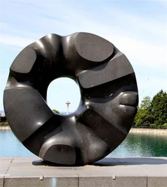 Large, abstract black sculpture in front of a body of water with trees and a distant tower.