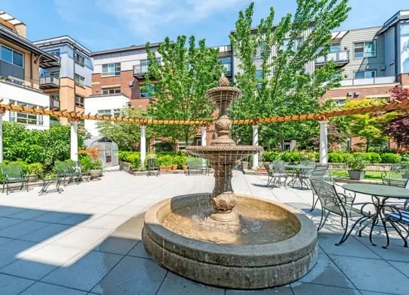 Exterior community courtyard with a central fountain, patio tables, and green trees.