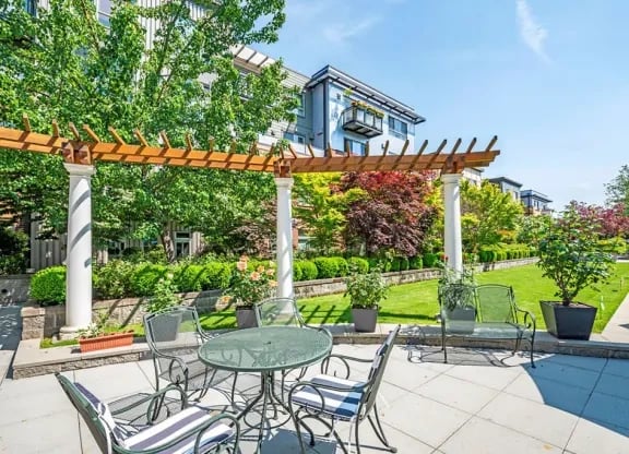 Outdoor community courtyard with a wooden pergola, round metal tables, and chairs amid manicured greenery.