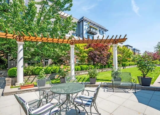 Outdoor communal patio with a round glass table and metal chairs under a wooden pergola, surrounded by trees.