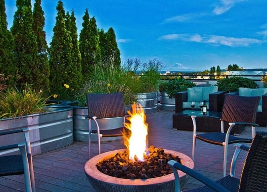 Outdoor rooftop lounge with a fire pit, wicker chairs, and planters on a wooden deck under a blue sky.