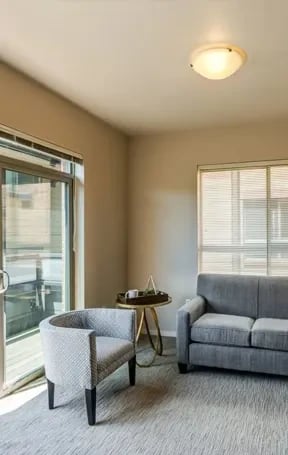 Living room with a gray sofa, patterned armchair, round side table, and a sliding glass door to a balcony.