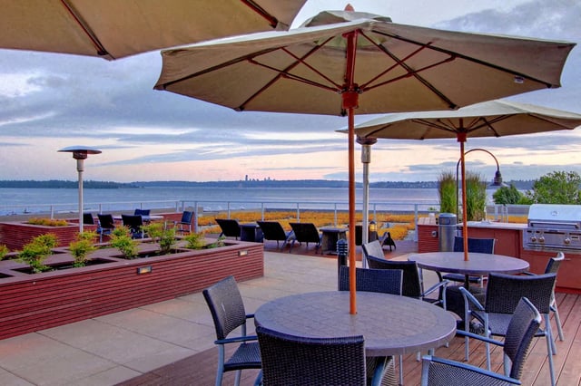 Rooftop patio with tables, chairs, umbrellas, and a grill overlooking the water at sunset.