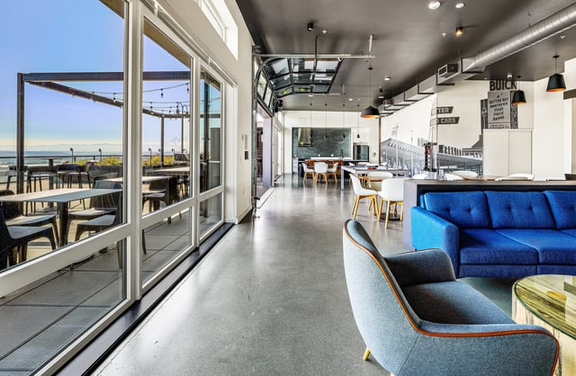 Modern communal lounge area with large windows, seating, and tables, leading to an outdoor patio with a view.